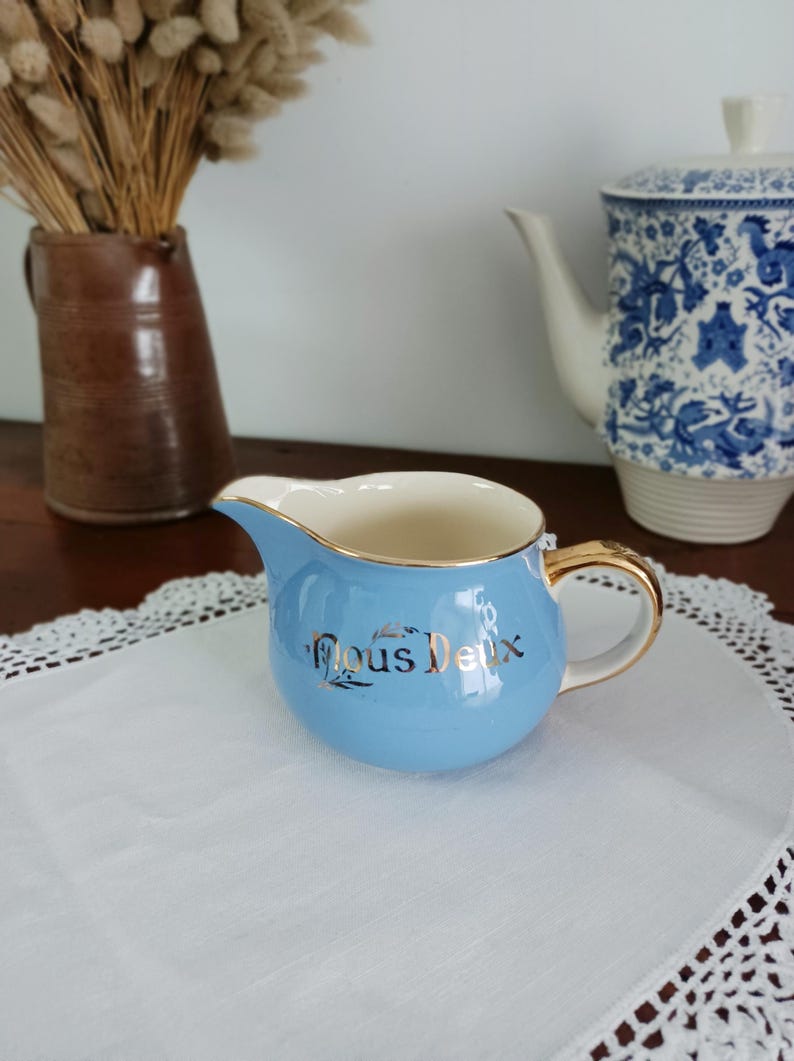 May include: A light blue creamer with a gold rim and handle. The creamer has a gold inscription that reads "Nous Deux".