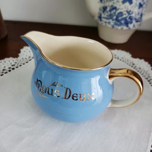May include: A light blue creamer with a gold rim and handle. The creamer has the words "Nous Deux" written in gold on the front.