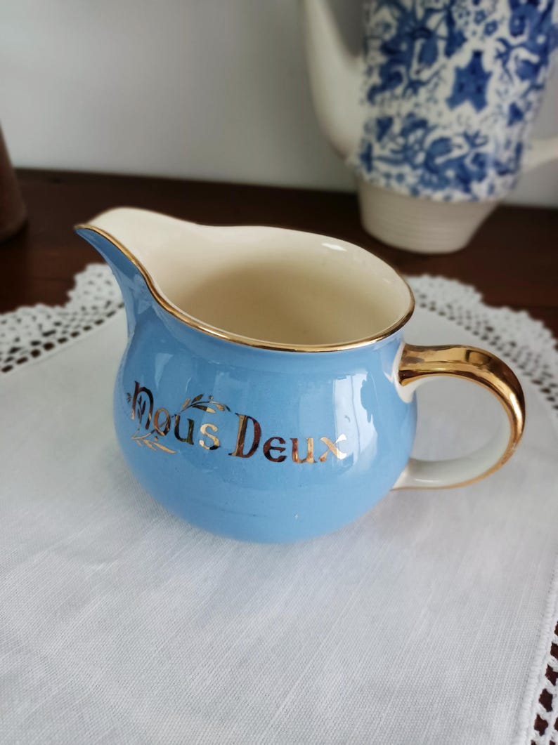May include: A light blue creamer with a gold rim and handle. The creamer has the words "Nous Deux" written in gold on the front.