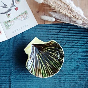 Op de afbeelding: Een keramische schelpvormige schaal met een gele rand en een bruin en groen interieur. De schaal staat op een blauwgroen oppervlak, met een vintage boek en gedroogde bloemen op de achtergrond. De schaal is waarschijnlijk voor decoratieve doeleinden.