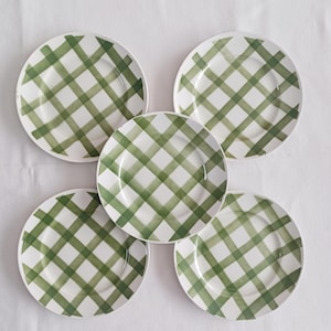 May include: Five white ceramic plates with a green plaid pattern. The plates are arranged in a circular pattern on a white surface. The green plaid design is made of intersecting lines.