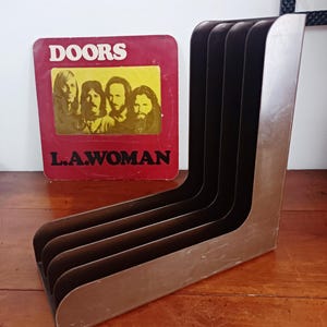 Puede incluir: Un estante de discos de metal con cinco ranuras para guardar discos de vinilo. El estante es de color marrón oscuro y tiene un diseño ligeramente curvado. Una portada de álbum de vinilo vintage para "The Doors" es visible en el fondo.