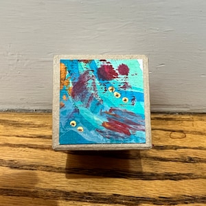 May include: A small, square, wooden box with a colorful abstract painting on the top. The painting features shades of blue, purple, and red with gold accents.