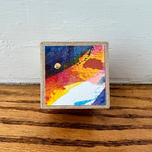 May include: A small, square box with a colorful abstract design on the front. The design features a mix of blue, yellow, orange, red, and white colors. There is a small gold circle in the upper left corner of the design.