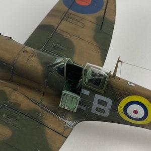 May include: A detailed model of a green and brown camouflage painted Supermarine Spitfire fighter plane with a partially open cockpit. The plane has a roundel on the side with the letters "EBOC" painted on the fuselage.