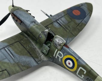 May include: A detailed model of a Supermarine Spitfire fighter plane with a green, blue, and gray camouflage paint scheme. The plane has a roundel on the wing and the letters "IROC" on the side. The cockpit is open and the pilot's seat is visible.