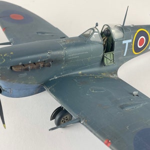 May include: A detailed model of a gray and blue Supermarine Spitfire fighter plane with a red, white, and blue roundel on the side. The plane has the letters "TOL" and the number "EP706" painted on the side.