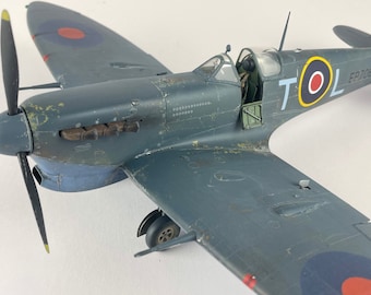 May include: A detailed model of a gray and blue Supermarine Spitfire fighter plane with a red, white, and blue roundel on the side. The plane has the letters "TOL" and the number "EP706" painted on the side.