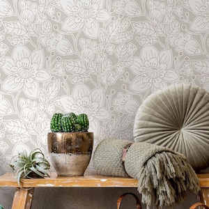 May include: A neutral-toned room with floral wallpaper and a wooden bench. The bench holds a potted cactus, a small succulent, and decorative pillows. A woven basket and a fringed throw add texture to the scene.