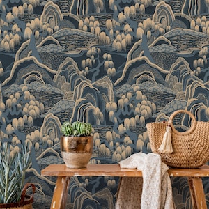 Op de afbeelding: Een kamer met behang met een blauw en goud berglandschap. Op een houten bank staan een geweven handtas met een kwast, een beige deken, een stapel boeken en een cactus in een pot. Een geweven mand met een plant staat op de vloer.