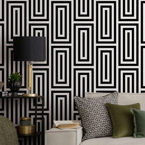 Op de afbeelding: Een woonkamer met een crèmekleurige bank en groene en zwarte kussens. Een zwart-gouden lamp staat op een bijzettafel. De muur is versierd met een zwart-wit geometrisch patroon.