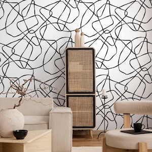 May include: A modern living room with a white wall featuring a black abstract line design. The room includes a beige sofa, wooden coffee table, woven cabinet, and a beige chair. Decorative items include a vase, black bowl, and a small black cup.