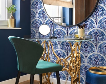 May include: A gold metal vanity table with a glass top and ornate legs. The table is set against a white and blue patterned wallpaper. A green velvet chair sits next to the table.