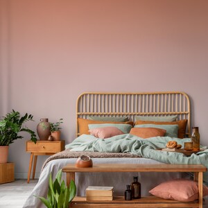 May include: A bedroom scene with a rattan headboard, featuring a bed with sage green bedding, orange and pink pillows, and a wooden bench at the foot. A wooden nightstand with a vase and plants is on the left. The wall has a gradient of pink and peach.