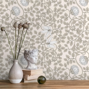 Op de afbeelding: Behang met beige bloemen met een herhalend patroon van gestileerde bloemen en bladeren in grijstinten en beige. Een vaas met gedroogde zaaddozen, een buste, boeken en een decoratieve glazen bol staan op een houten oppervlak.