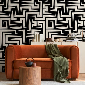 May include: A burnt orange velvet sofa with a green throw blanket in front of a black and white geometric patterned wall. A small wooden table with a black teapot and a saucer sits in front of the sofa.