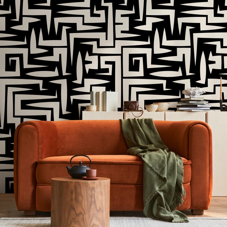 May include: A burnt orange velvet sofa with a green throw blanket in front of a black and white geometric patterned wall. A small wooden table with a black teapot and a saucer sits in front of the sofa.
