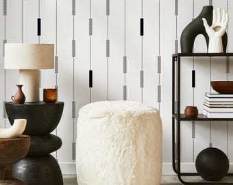 May include: A room scene featuring a white, fluffy ottoman, a black side table with a lamp, and a black metal shelf. The background is a white wall with a black and gray linear pattern. Decorative items include vases and a sculpture.