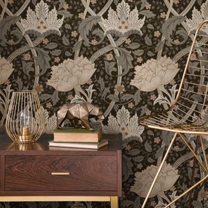 May include: A dark brown floral wallpaper with large cream and gray flowers and leaves. A gold wireframe lamp, a geometric bear sculpture, and a gold chair are in front of the wallpaper. A dark wood nightstand with a drawer is in the foreground.