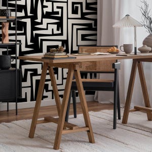 May include: A wooden trestle desk with a black woven chair in front of it. The desk has a white and black geometric patterned wall behind it.