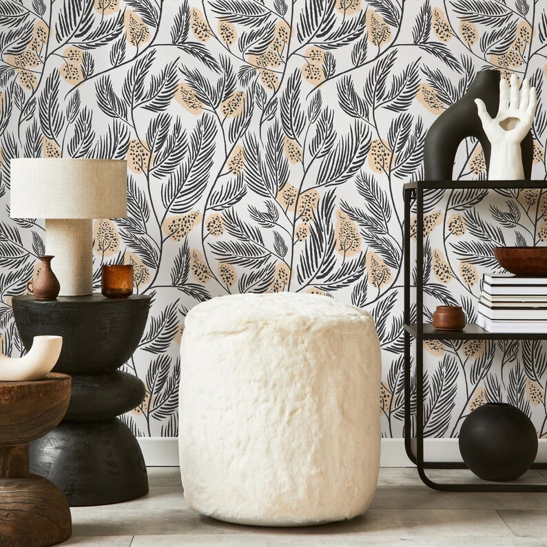 May include: A white faux fur ottoman with a black and beige floral wallpaper background. The ottoman is sitting in front of a black metal shelf with a bowl and books on it.