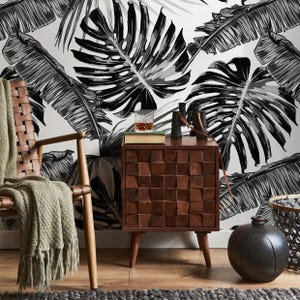 May include: A wooden nightstand with a geometric design on the drawer front, featuring a glass of whiskey, binoculars, and a book. The background is a black and white tropical leaf wallpaper. A woven chair and a green blanket are in the foreground.
