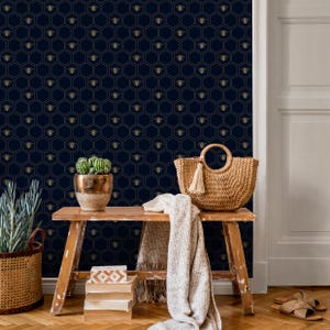May include: A navy blue wallpaper with a repeating pattern of gold bees within honeycomb shapes. A wooden bench holds a woven tote bag with a tassel, a potted cactus, and a folded blanket. A wicker basket and a stack of books are also visible.