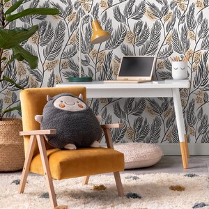 May include: A yellow velvet armchair with a gray plush monster pillow. The chair is in front of a white desk with a laptop and a white lamp with a yellow shade. The wall behind the desk has a black and white floral wallpaper.