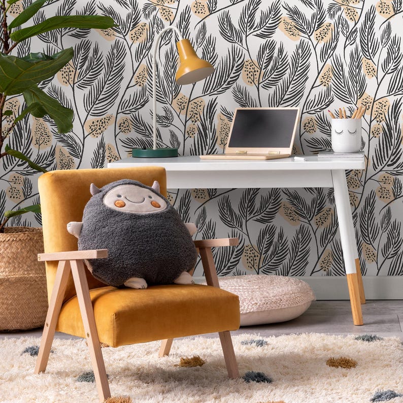 May include: A yellow velvet armchair with a gray plush monster pillow. The chair is in front of a white desk with a laptop and a white lamp with a yellow shade. The wall behind the desk has a black and white floral wallpaper.