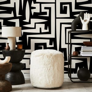 May include: White faux fur ottoman with a black and white geometric patterned wall behind it. The ottoman is round and sits on a wooden floor.