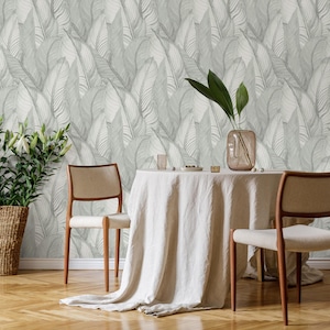 May include: A dining room setting with a round table covered by a white linen tablecloth. Two wooden chairs with beige seats are placed around the table. The backdrop is a wallpaper with a large, repeating leaf pattern in grey and white tones.