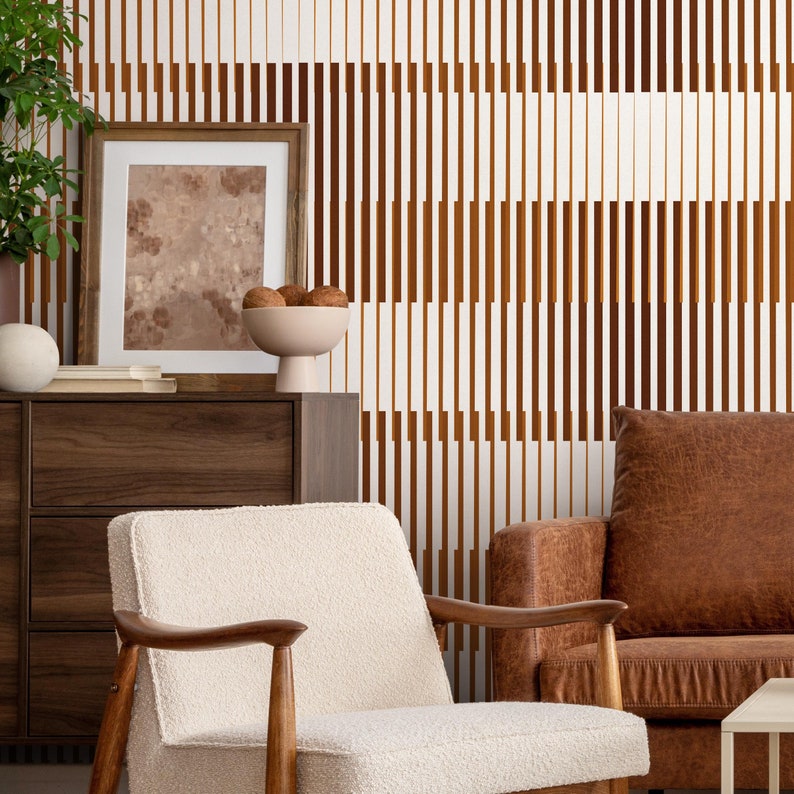 Op de afbeelding: Een woonkamerinterieur met een witte muur met een bruin-wit geometrisch patroon behang. Een witte fauteuil met houten poten staat voor een bruine leren bank. Een houten dressoir met twee lades staat aan de linkerkant van de afbeelding. Een ingelijst schilderij hangt aan de muur boven het dressoir.