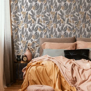 May include: A bedroom with a bed featuring a peach-colored duvet and a yellow throw blanket. The bed has a gray pillow and a black pillow. The wall behind the bed is covered in a white wallpaper with a black and yellow floral pattern.