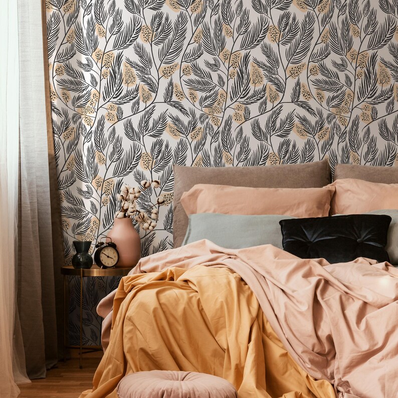 May include: A bedroom with a bed featuring a peach-colored duvet and a yellow throw blanket. The bed has a gray pillow and a black pillow. The wall behind the bed is covered in a white wallpaper with a black and yellow floral pattern.