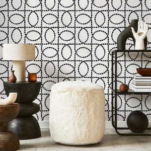 May include: A modern interior scene featuring a white, fluffy ottoman, a black side table with a lamp, and a black metal shelf. The background is a white wall with a black oval pattern. Decorative items are on the table and shelf.
