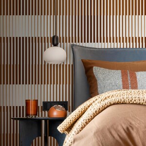 Op de afbeelding: Een slaapkamer met een bruin-wit gestreepte muur, een bed met een bruin dekbed, een grijze deken en een bruin kussen met een grijs-oranje gestreept patroon. Een witte bolvormige hanglamp hangt boven een nachtkastje met een bruine klok en een glazen kaarsenhouder.