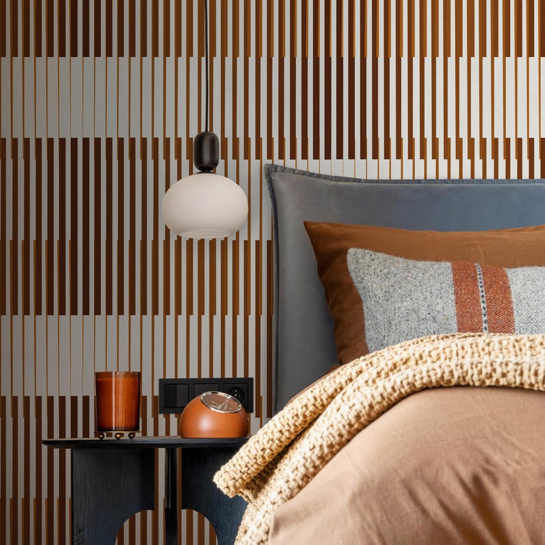 Op de afbeelding: Een slaapkamer met een bruin-wit gestreepte muur, een bed met een bruin dekbed, een grijze deken en een bruin kussen met een grijs-oranje gestreept patroon. Een witte bolvormige hanglamp hangt boven een nachtkastje met een bruine klok en een glazen kaarsenhouder.