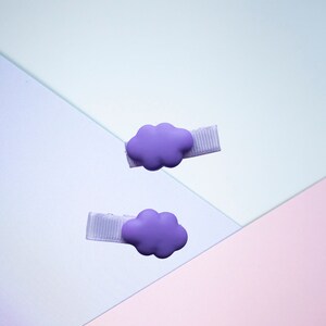 Op de afbeelding: Twee paarse haarclips in de vorm van een wolk met witte lintaccenten. De clips bevinden zich op een lichtblauwe en roze achtergrond.