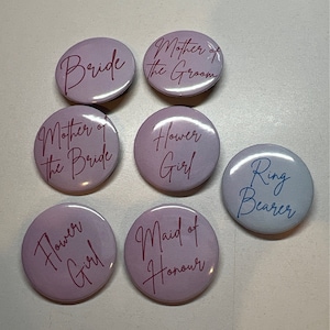 May include: Eight round buttons with different wedding party titles printed on them. The buttons are in shades of purple and blue with white text. The titles include "Bride", "Mother of the Bride", "Mother of the Groom", "Maid of Honour", "Flower Girl", "Flower Girl", and "Ring Bearer".