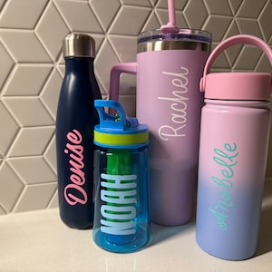 May include: A collection of personalized water bottles and tumblers. Includes a navy blue bottle with "Denise" in pink, a blue bottle with "Noah" in white, a purple tumbler with "Rachel" in white, and a pink and blue ombre bottle with "Anabelle" in teal.