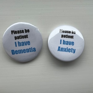 Peut inclure: Deux boutons ronds blancs avec du texte bleu et noir. Le bouton de gauche dit "Veuillez être patient J'ai la démence". Le bouton de droite dit "Veuillez être patient J'ai de l'anxiété".