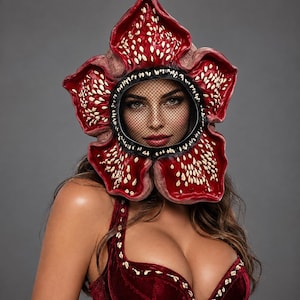 Peut inclure: Un masque intégral en forme de fleur épanouie avec cinq grands pétales rouge foncé. Le centre présente un filet noir couvrant la zone du visage. Les pétales sont texturés avec de nombreuses petites protubérances blanc cassé en forme de dents.