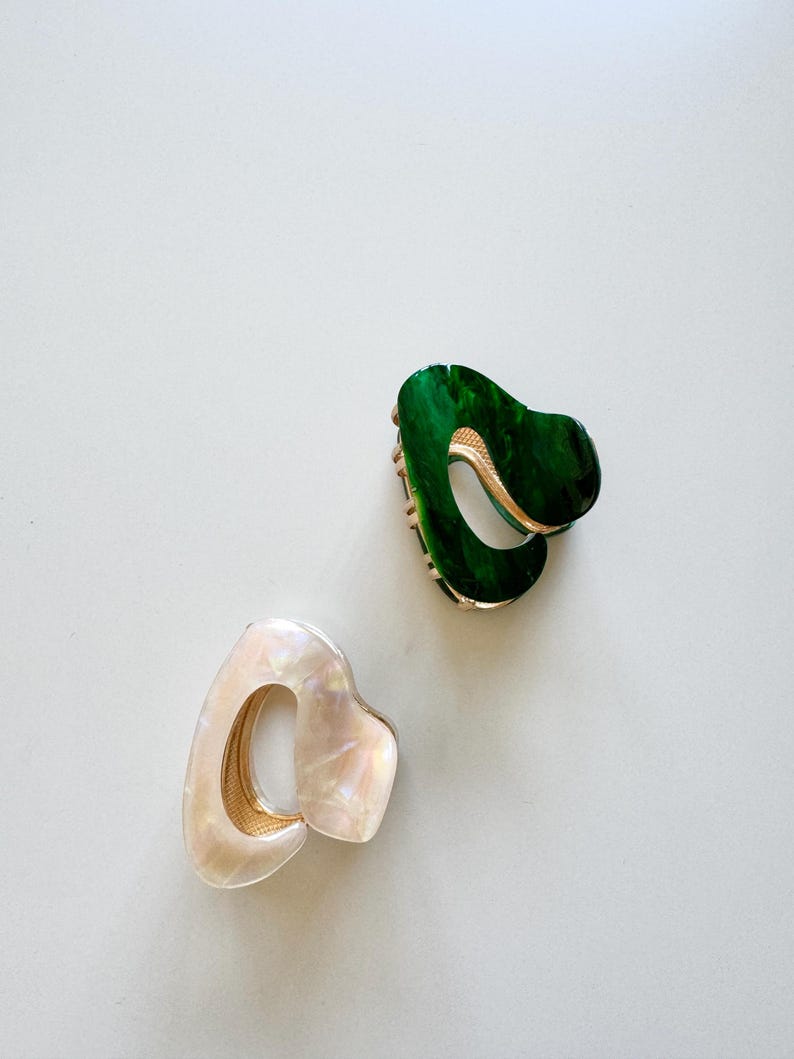 May include: Two hair clips, one green and one white, with a gold metal accent. The clips are made of a textured plastic material.