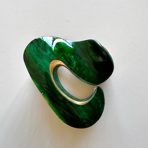 May include: A green and gold abstract hair clip. The clip is made of a smooth, shiny material and has a unique, organic shape.