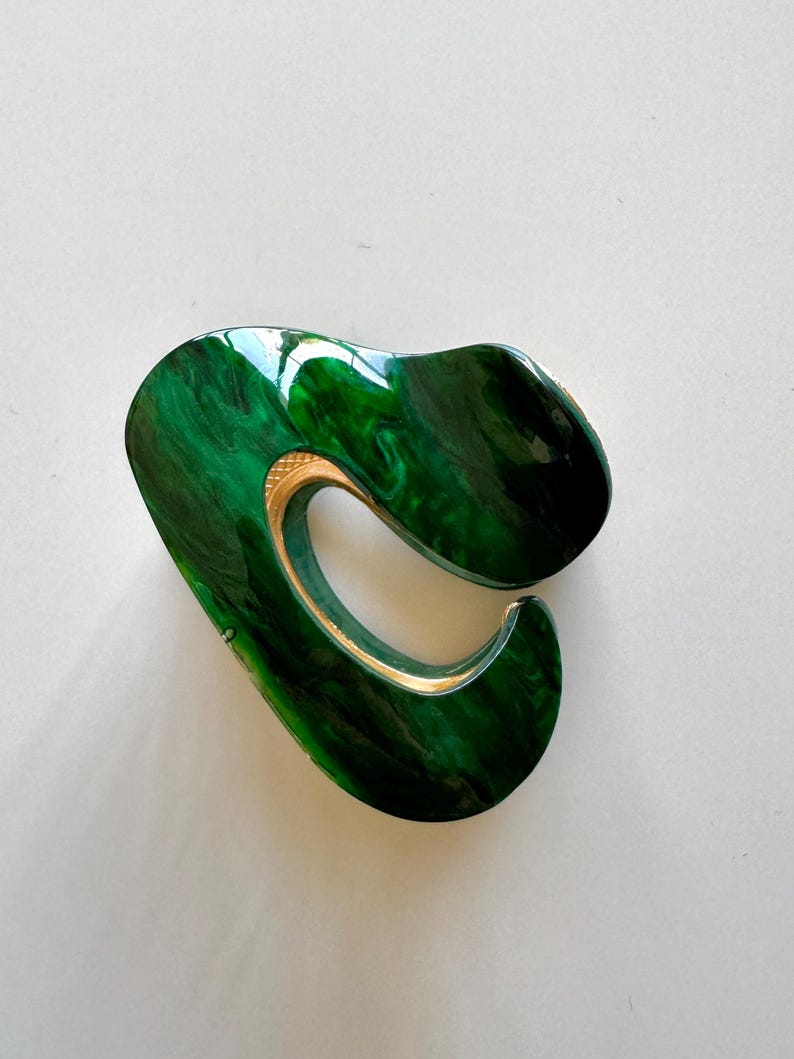 May include: A green and gold abstract hair clip. The clip is made of a smooth, shiny material and has a unique, organic shape.