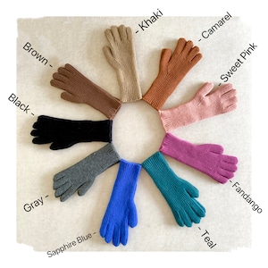 Op de afbeelding: Tien paar lange gebreide handschoenen in verschillende kleuren, gerangschikt in een cirkel op een licht beige oppervlak. De kleuren zijn: bruin, zwart, grijs, saffierblauw, teal, fandango, roze, camel, kaki.