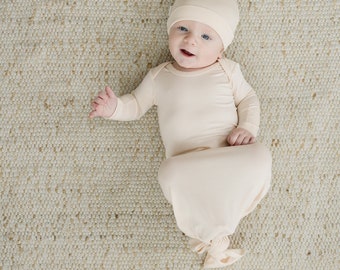 Könnte beinhalten: Ein Baby trägt ein hellbeiges, geknotetes Kleid und eine passende Mütze. Das Baby liegt auf einem weißen, strukturierten Teppich.