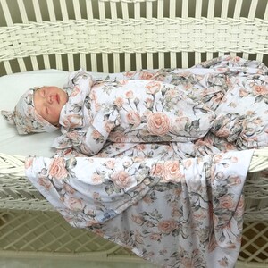 May include: A baby is sleeping in a white wicker bassinet. The baby is wearing a white and pink floral swaddle and a matching hat. The swaddle and hat have a floral pattern with pink roses and green leaves.