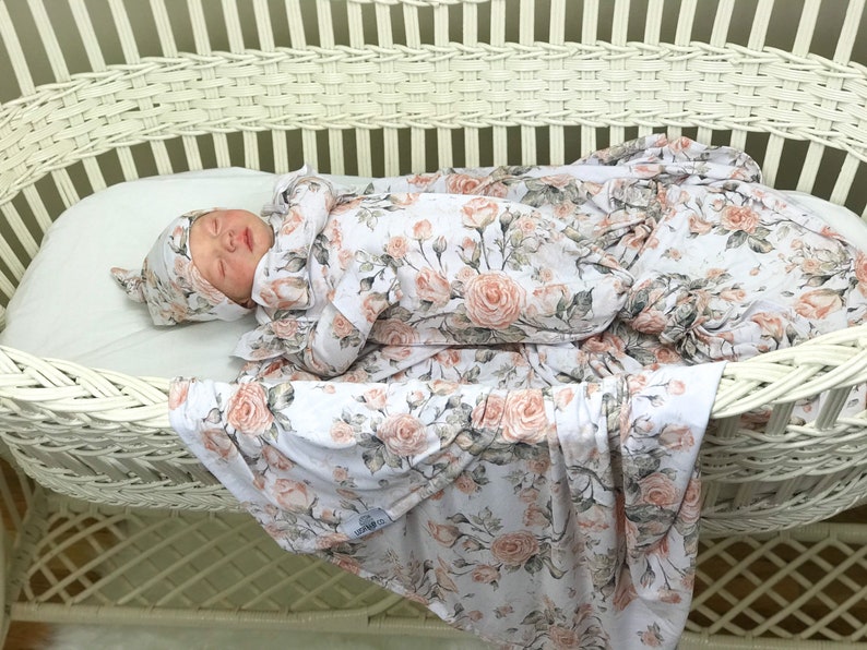 May include: A baby is sleeping in a white wicker bassinet. The baby is wearing a white and pink floral swaddle and a matching hat. The swaddle and hat have a floral pattern with pink roses and green leaves.
