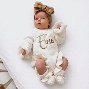 May include: An off-white long-sleeve knit romper featuring a ruffled hem and cuffs, with the name Evie embroidered in brown script across the chest. The outfit is paired with a matching large brown fabric bow headband and knit booties.